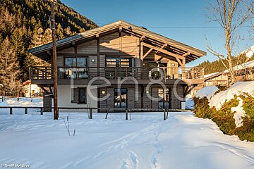 A large wooden chalet with balconies is set in deep snow against a backdrop of a forested mountain under a clear blue winter sky.