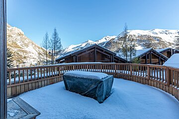 A pristine snowy wooden terrace with a covered outdoor grill, overlooking rustic chalets and majestic snow-capped mountains under a clear blue sky.