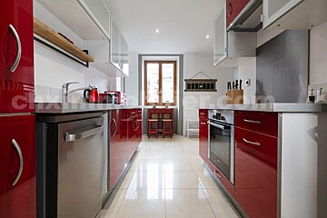 Sleek galley kitchen with bright red cabinets, white uppers, and stainless steel appliances. A window and small red table sit at the far end.