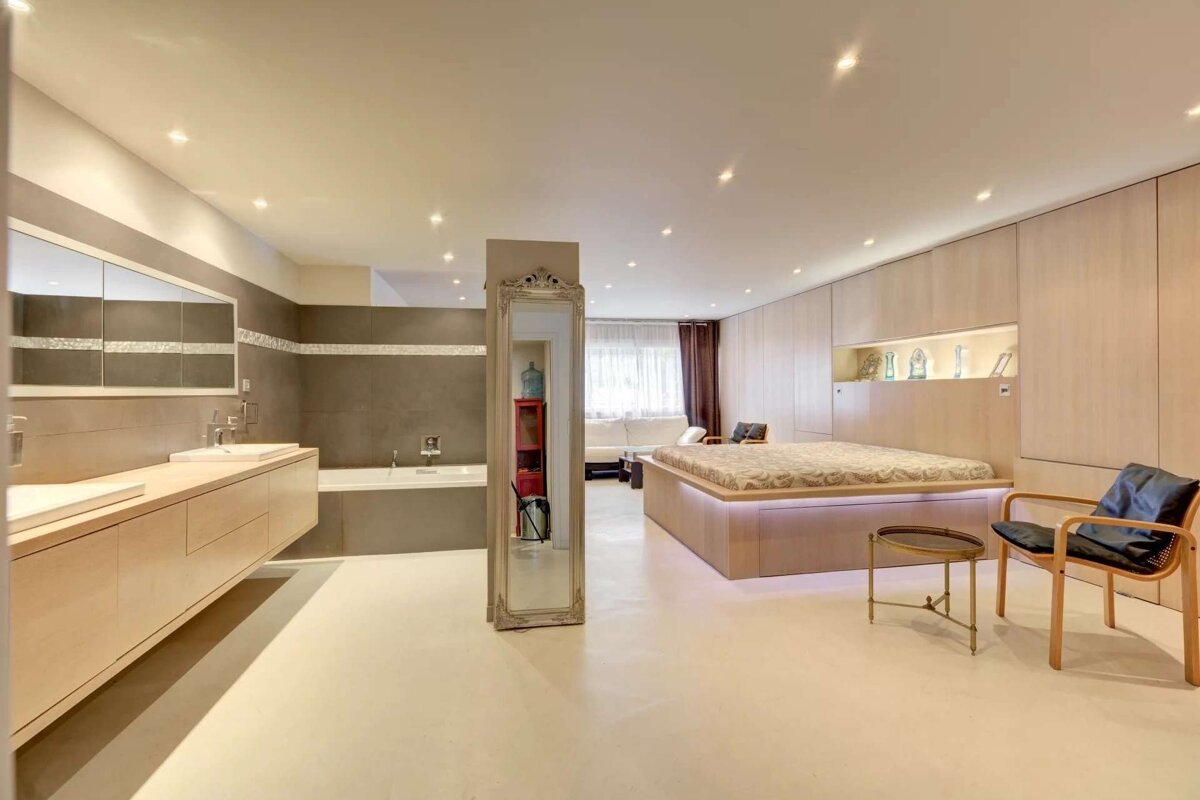 Chic open-plan bedroom and bathroom with light wood and grey accents. Features a modern bed, double vanity, and ornate mirror divider.
