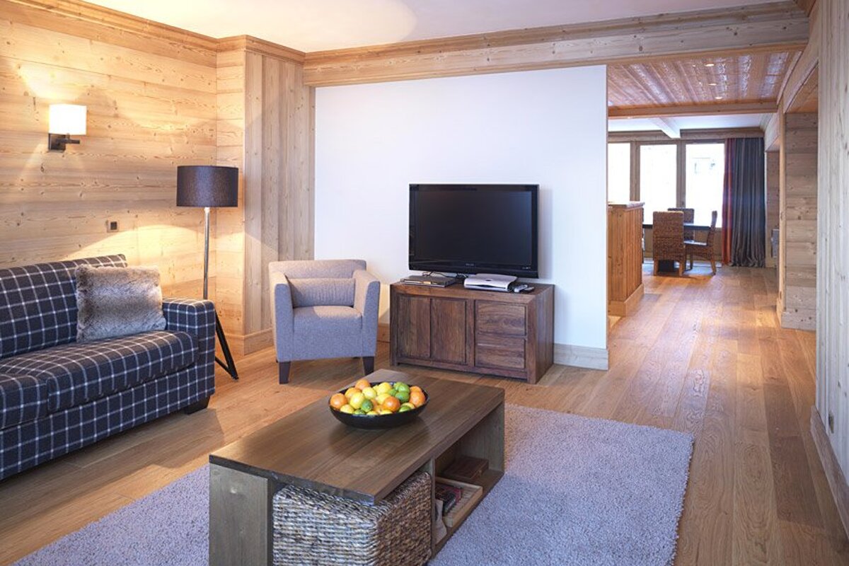 A cozy, wood-paneled living room with a plaid sofa, grey armchair, coffee table with fruit, TV, and light rug, open to a dining area.