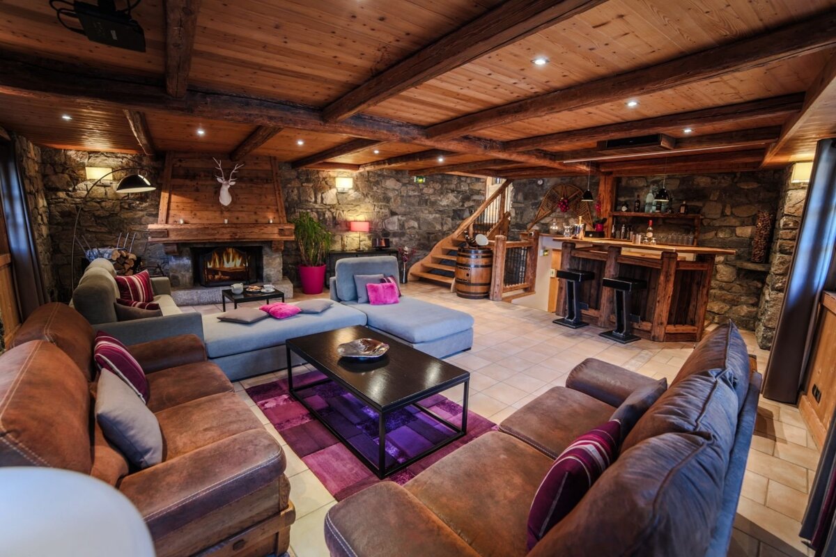 A cozy, rustic chalet living room with stone walls, wood ceilings, a fireplace, plush seating, and a bar, all warmly lit with pops of pink/purple.