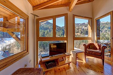 A cozy, wood-accented room with large windows offers panoramic views of a sunny, snow-dusted mountain forest. It features a TV and a leather armchair.