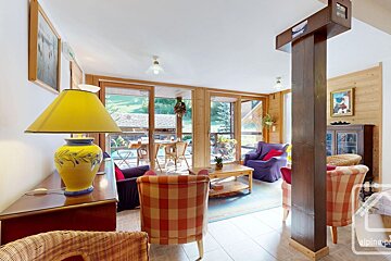 A bright, rustic living room with large glass doors opening to a green view. Features plaid armchairs, wooden accents, and a yellow lamp.