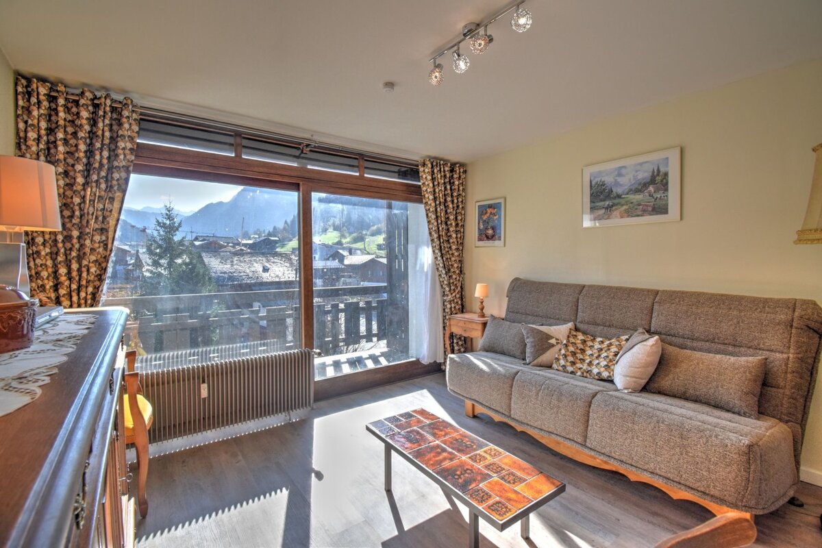 A bright living room features a sofa, tiled coffee table, and large sliding doors opening to a balcony with stunning mountain and village views.