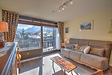 Sunlit living room with a sofa, coffee table, and large sliding doors leading to a balcony with a picturesque mountain village view.