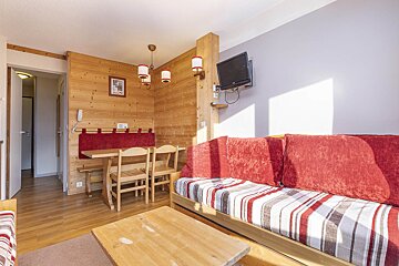 Cozy chalet-style living room with wood paneling, a red-cushioned sofa, wooden dining set, and a wall-mounted TV.