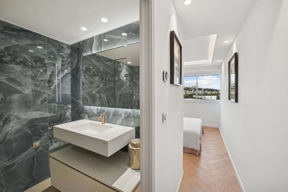 A modern bathroom with grey marble walls and a white floating sink, adjacent to a hallway leading to a bright bedroom with a large window view.