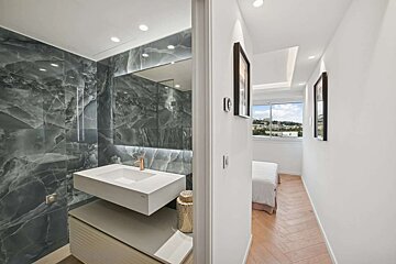 A modern bathroom with grey marble walls and a white floating sink, adjacent to a hallway leading to a bright bedroom with a large window view.