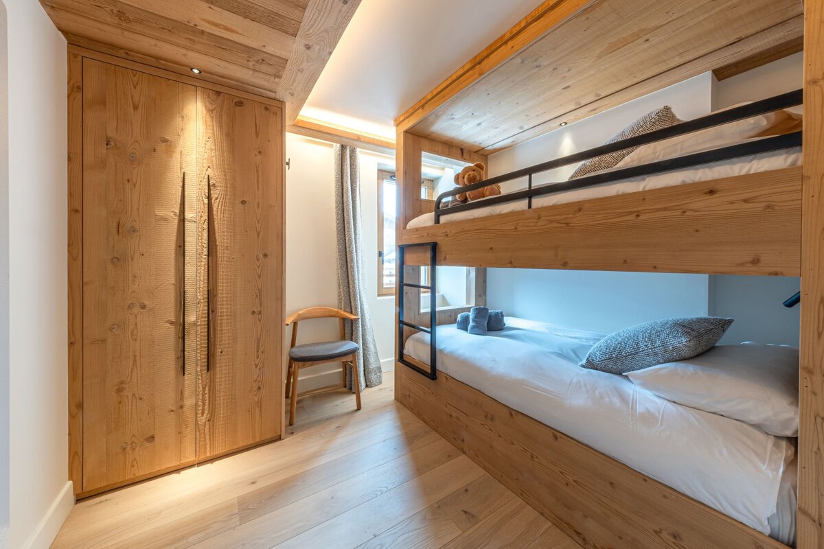 A modern rustic bedroom featuring light wood bunk beds, a built-in wardrobe, and light wood flooring. A cozy space with a window and small chair.