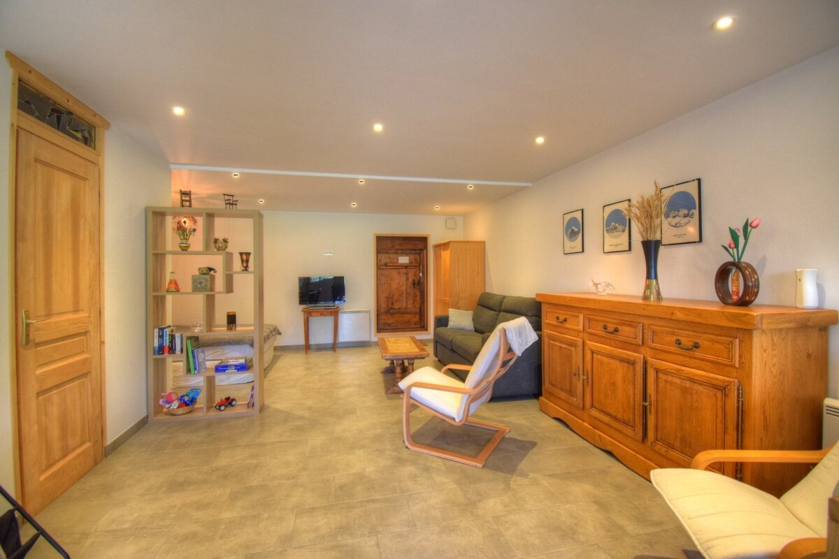 Bright living room featuring light wood furniture, a grey sofa, modern armchairs, tile flooring, and warm recessed lighting.