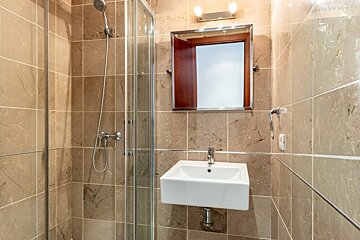 A modern bathroom features beige tiled walls, a glass shower, a white square sink, and a wooden-framed mirror with lights above it.