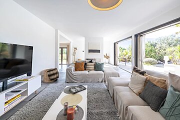 A bright, contemporary living room with neutral sofas, a TV, and large glass doors leading to a lush green garden.