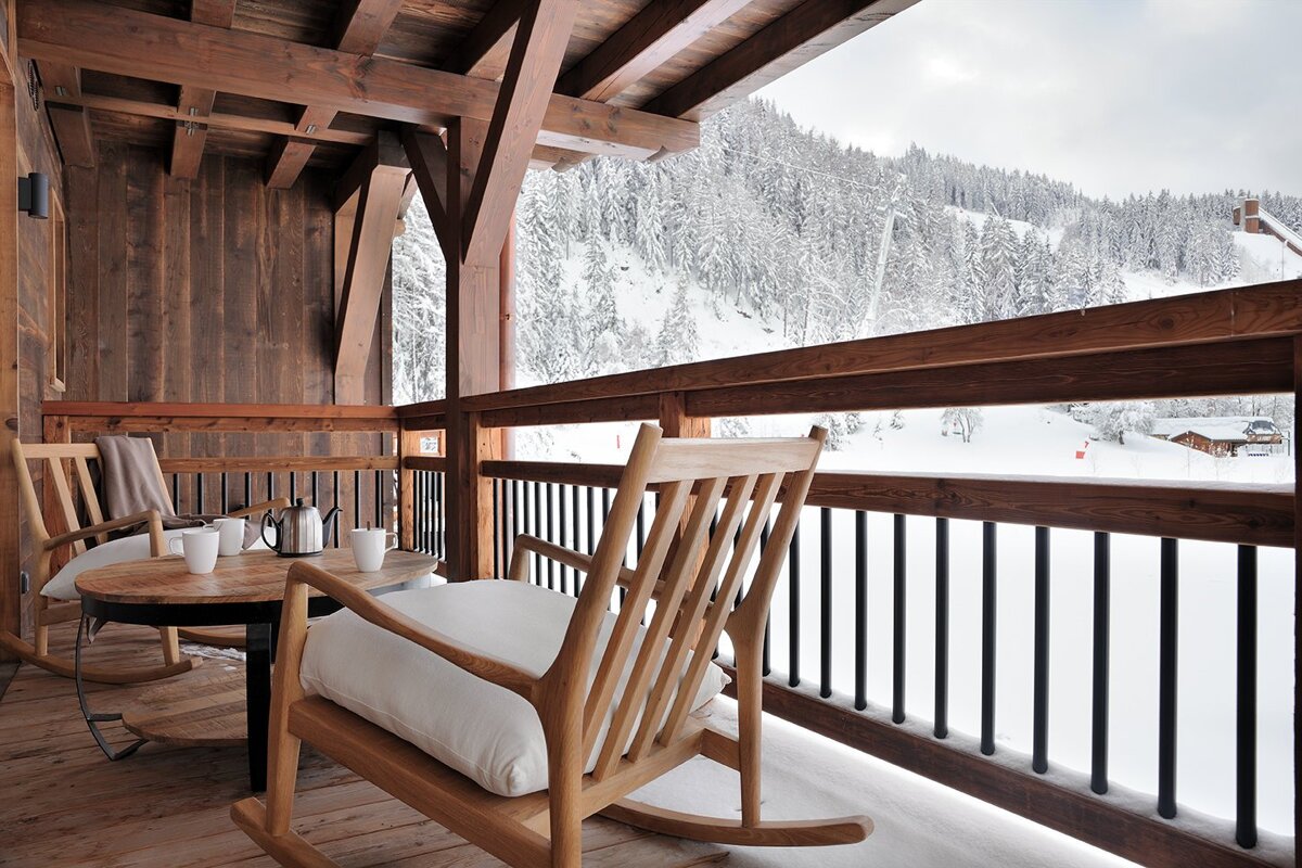 A cozy wooden balcony of an alpine chalet with rocking chairs and a coffee table overlooking a beautiful snowy forest.