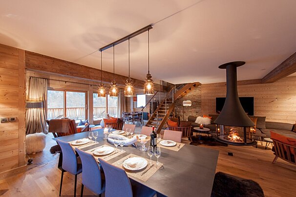 A spacious, modern chalet interior featuring a large dining table, a cozy living area with a suspended fireplace, and warm wooden accents throughout.