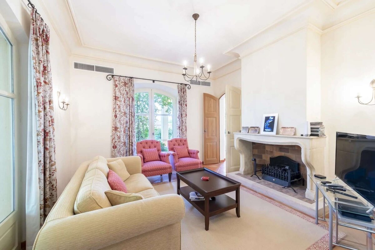 An inviting living room featuring a cream sofa, pink armchairs, dark coffee table, a grand fireplace, and a TV, with patterned curtains and a chandelier.