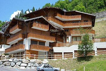 A large, multi-story wooden chalet-style building with many balconies on a green hillside, with a car parked nearby.