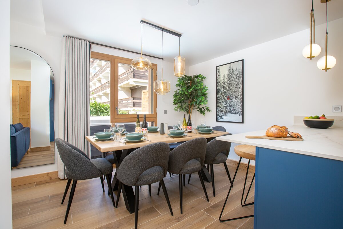 A stylish dining area features a wooden table set for dinner with grey chairs, modern pendant lights, and a window view. A kitchen island is to the right.