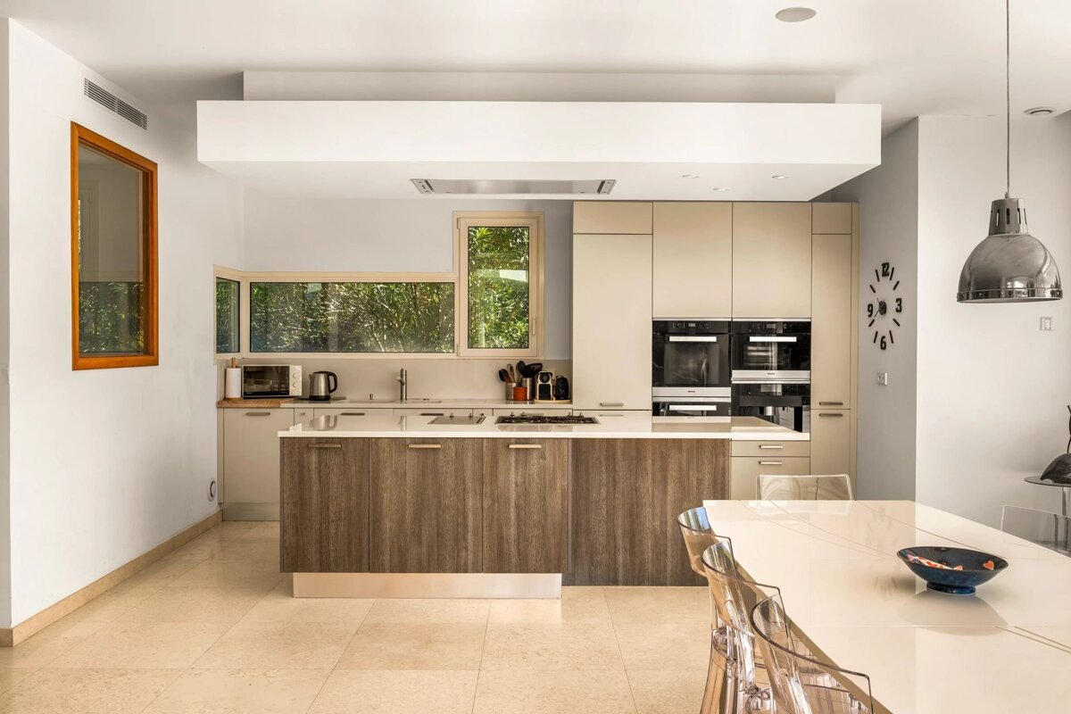 A bright, modern kitchen featuring wood and cream cabinets, built-in appliances, a large island, ample windows with greenery, and an adjacent dining area.