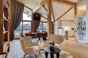 Modern chalet interior with exposed wood beams, large windows offering mountain views, and an open-plan living, dining, and kitchen area.