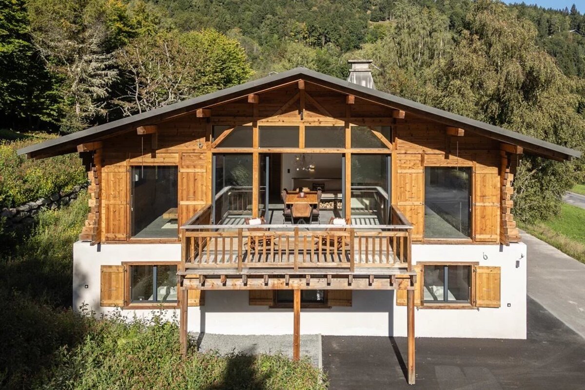 A two-story wooden chalet with a balcony, set against a backdrop of lush green forested hills.