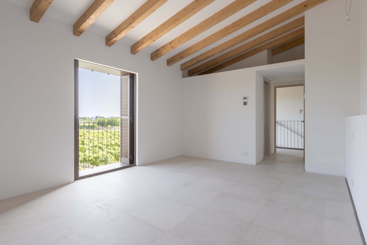 Bright, empty room with white walls, exposed wooden ceiling beams, and a large balcony opening to a lush green view. Light floor and an adjacent hallway.
