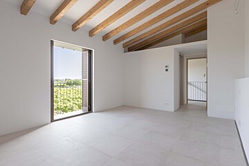 Bright, empty room with white walls, exposed wooden ceiling beams, and a large balcony opening to a lush green view. Light floor and an adjacent hallway.