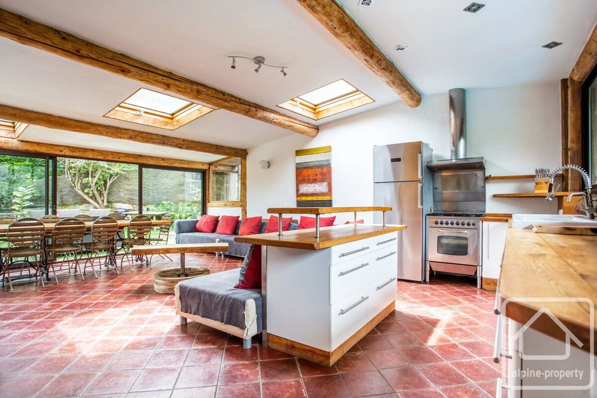 Modern rustic kitchen and living area with terracotta tiled floor, exposed wooden beams, skylights, and a central white island.