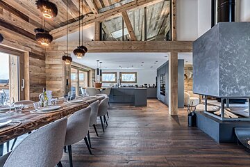 Luxurious open-plan chalet with a long wooden dining table, modern kitchen, contemporary fireplace, and rustic beamed ceiling.