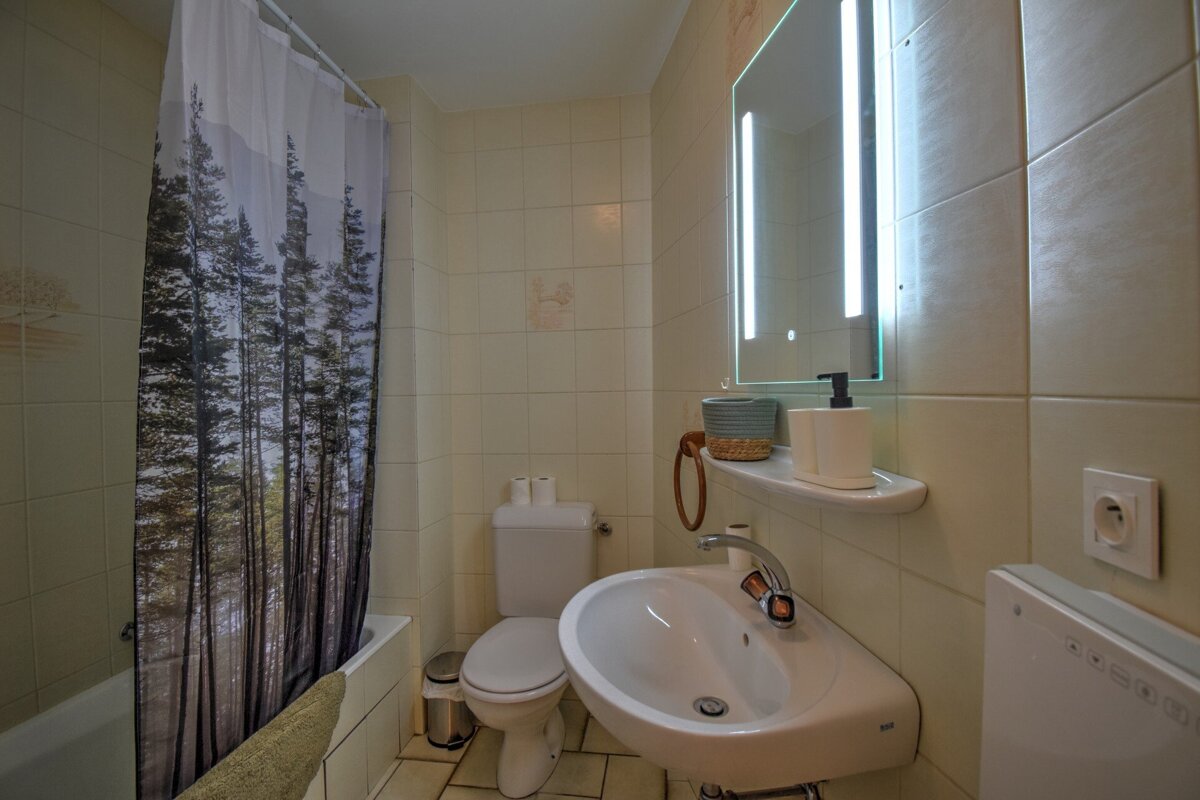 A cream-tiled bathroom features a forest-themed shower curtain, white sink, toilet, bathtub, and a backlit mirror above the vanity.
