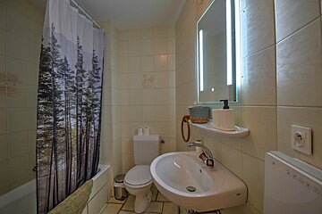A cream-tiled bathroom features a forest-themed shower curtain, white sink, toilet, bathtub, and a backlit mirror above the vanity.