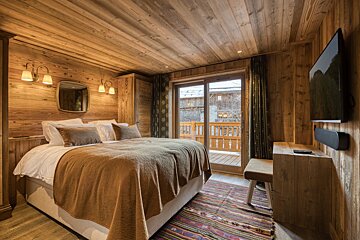 A cozy, wood-paneled bedroom features a comfy bed, balcony access to a snowy scene, and a wall-mounted TV.