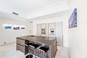 Sleek modern kitchen with a dark island and three bar stools, built-in ovens, and artwork on bright white walls.