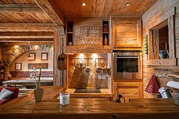 Cozy, all-wood chalet interior with a rustic kitchen featuring an oven, stovetop, hanging pots, and a glimpse of a dining area.