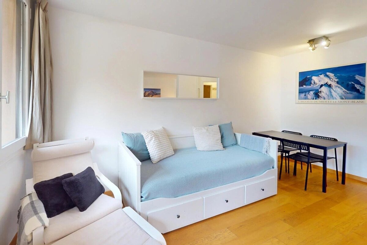 Bright room featuring a white daybed with light blue bedding, a cozy armchair, and a small dining set on wooden floors.