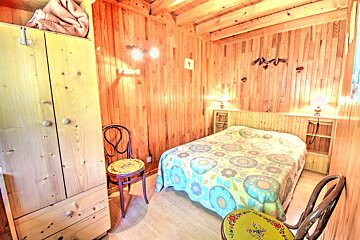 Cozy wooden room featuring a bed with a floral comforter, a light-colored wardrobe, and two decorative chairs. Warm lighting illuminates the space.