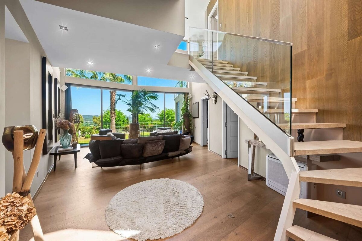 Bright, modern living room with floor-to-ceiling windows, palm tree views, wooden floor, and a sleek glass-railed staircase.