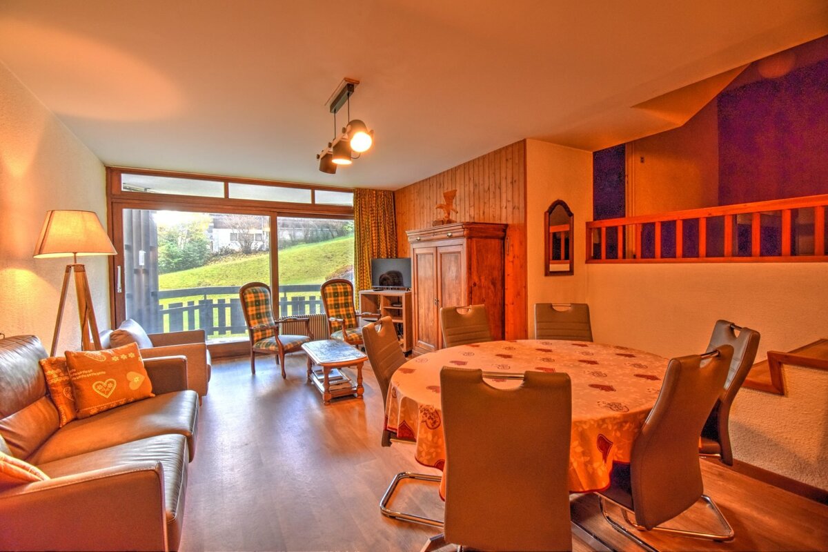 A cozy, warm-lit living and dining room with a large window overlooking a green landscape. Features a sofa, dining table, wooden cabinet, and indoor railing.