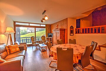 Cozy wooden chalet interior with a warm-toned living and dining area, featuring large windows looking out to a green hillside.