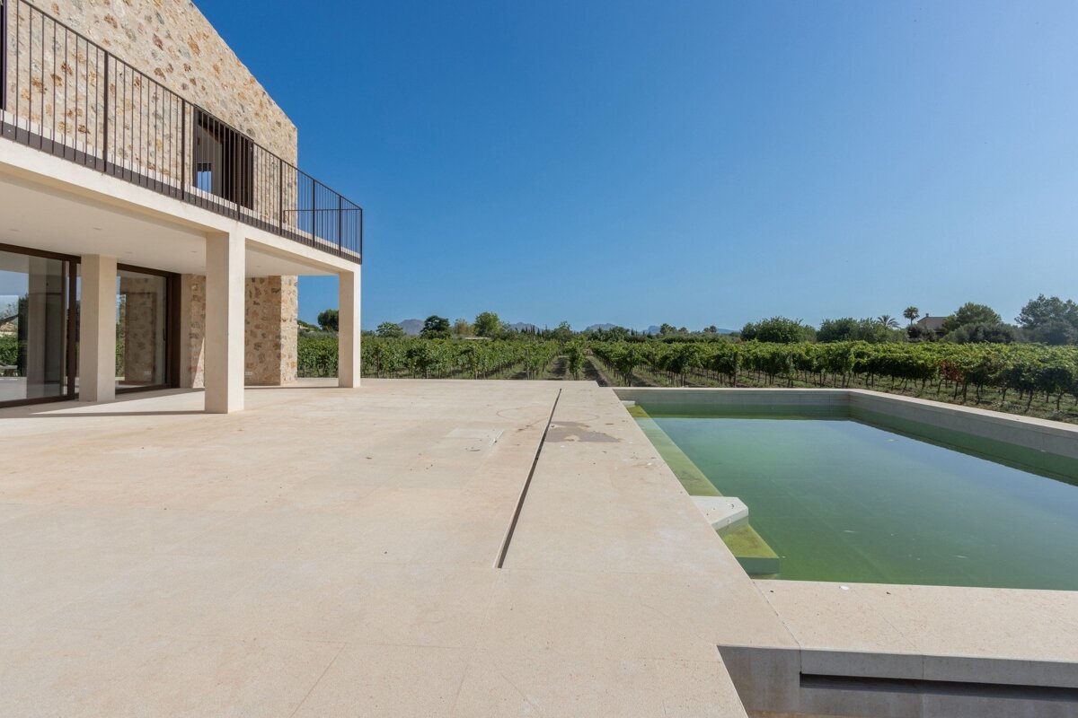 Modern stone villa with a patio, a rectangular green pool, and a vast vineyard under a clear blue sky.