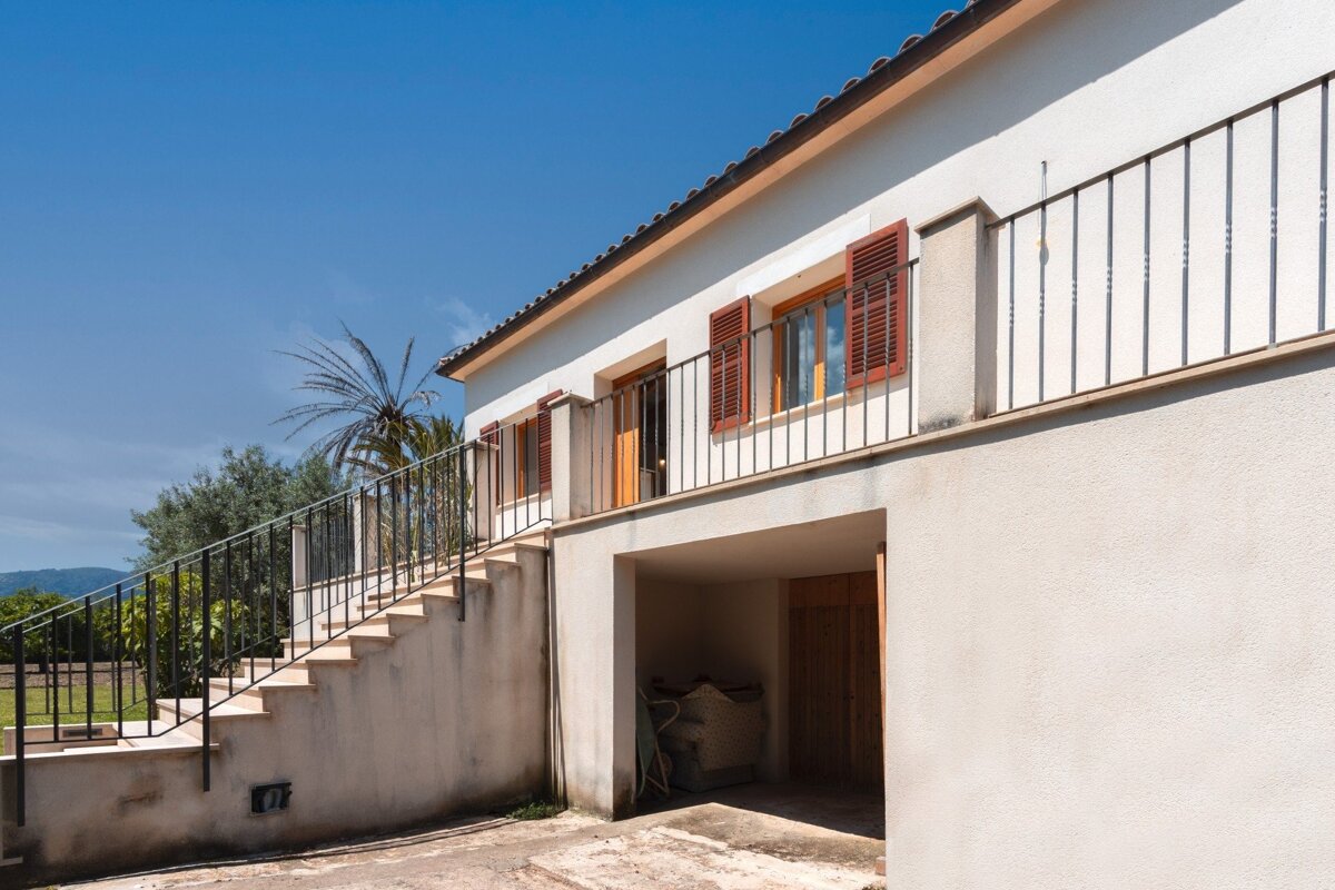 A white house with red-shuttered windows, a balcony with black railing, and stairs leading up. A garage is beneath, and a palm tree stands against a blue sky.