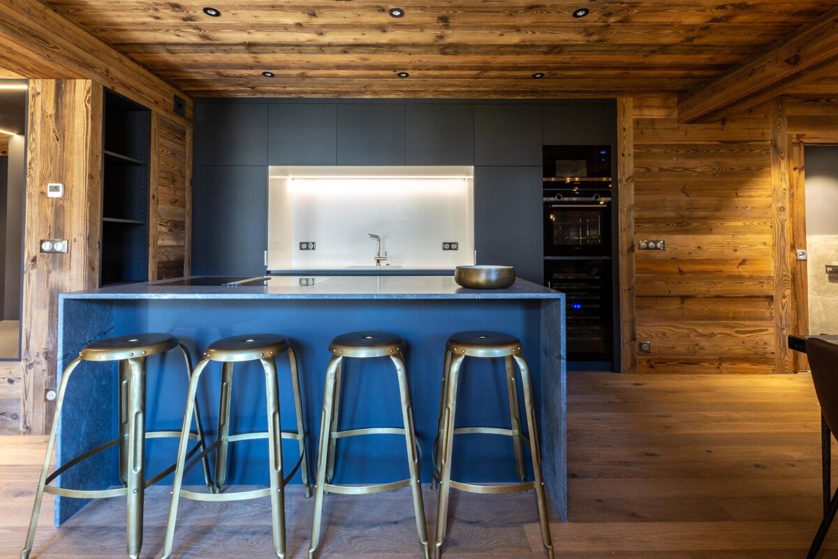 Modern kitchen in a rustic wooden cabin featuring a blue island with four brass stools and sleek dark cabinets.