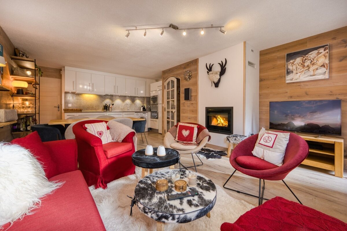 Cozy chalet-style living room with red seating, a lit fireplace, modern kitchen, and rustic wood accents. Cowhide rugs add warmth.