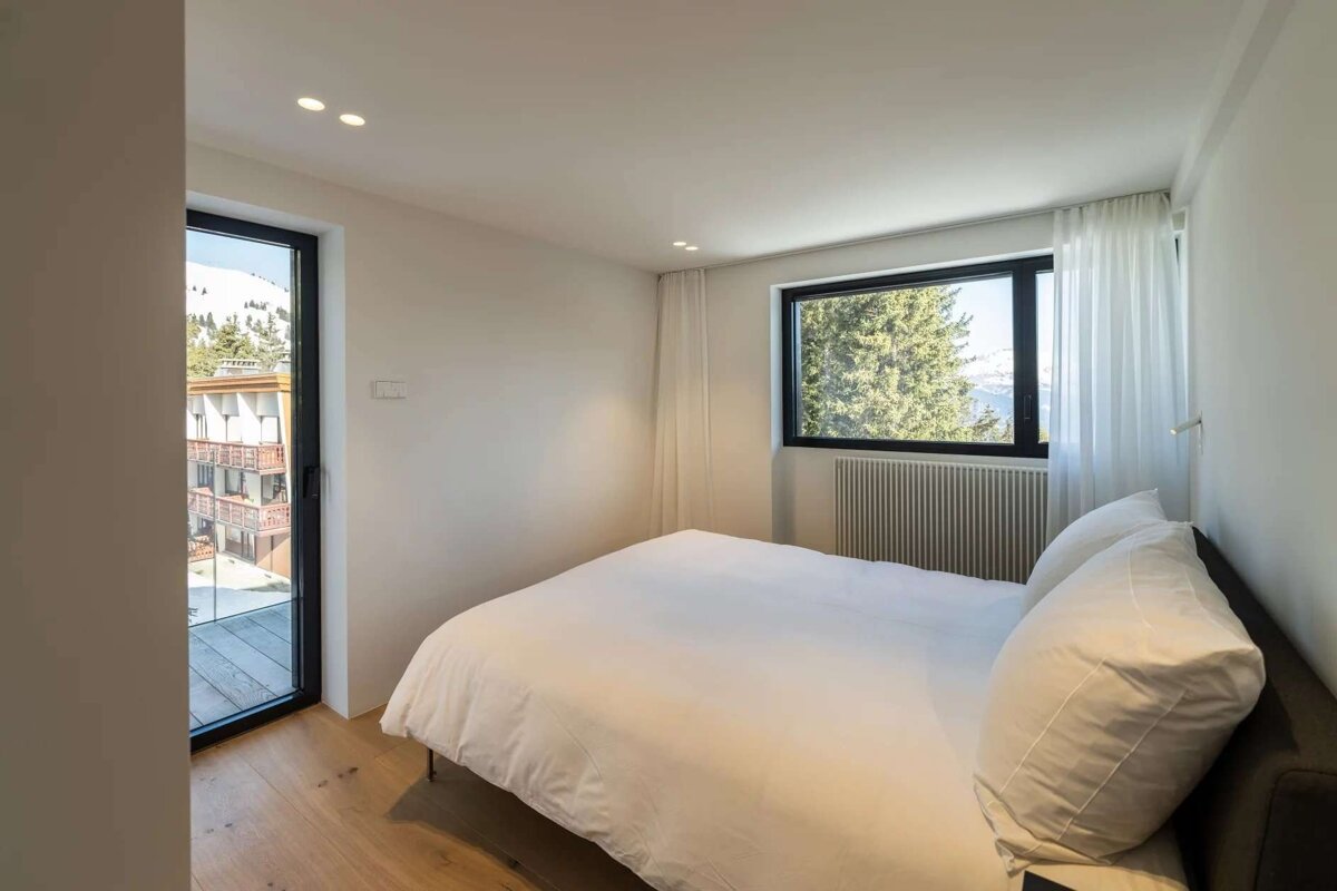 A modern, minimalist bedroom with white bedding, hardwood floors, and large windows offering views of mountains, trees, and a nearby building.