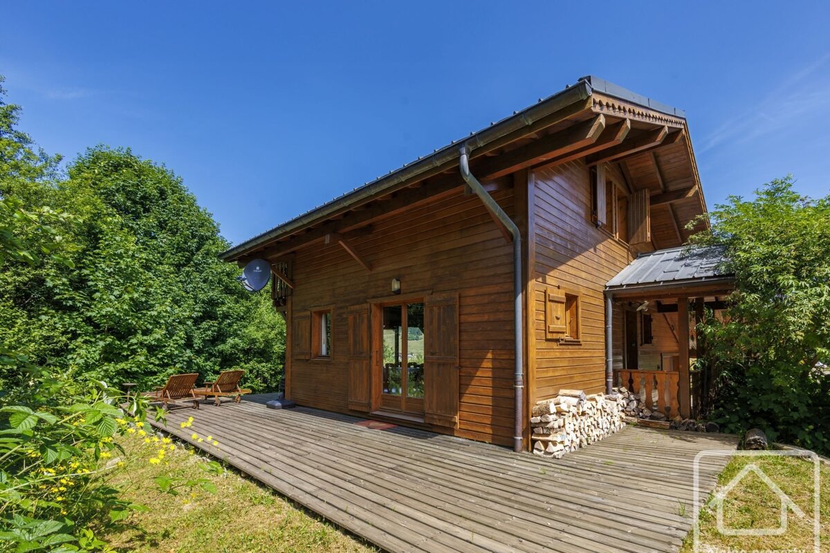 A rustic wooden house with a wide deck, surrounded by vibrant green foliage under a bright blue sky. Firewood is stacked neatly by the entrance.