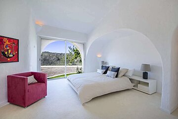 A serene, minimalist white bedroom with arched walls, a large window showcasing a green landscape, a white bed, and a vibrant red armchair.