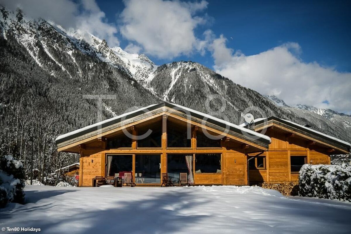 A cozy wooden chalet sits in a snowy landscape, framed by majestic, snow-capped mountains under a blue sky.