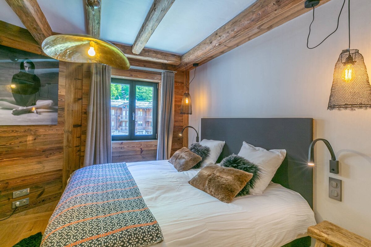Cozy chalet-style bedroom with warm wooden walls, exposed beams, and a plush bed. Unique lighting fixtures and a window view of buildings and trees.