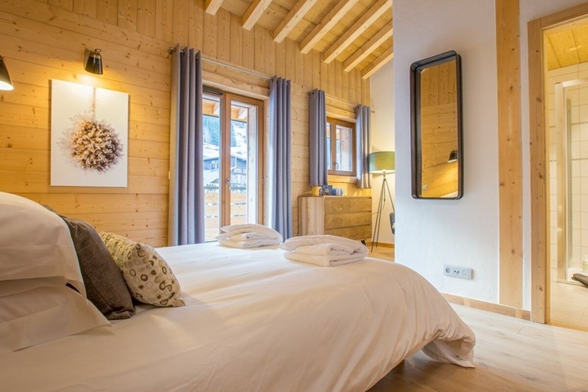 Cozy alpine chalet bedroom with warm wood paneling, a neatly made double bed, and glass doors leading outside.