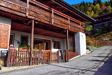 Cozy mountain house with a wooden balcony, fenced patio, and an elderly person. Autumn foliage covers the hills in the background.